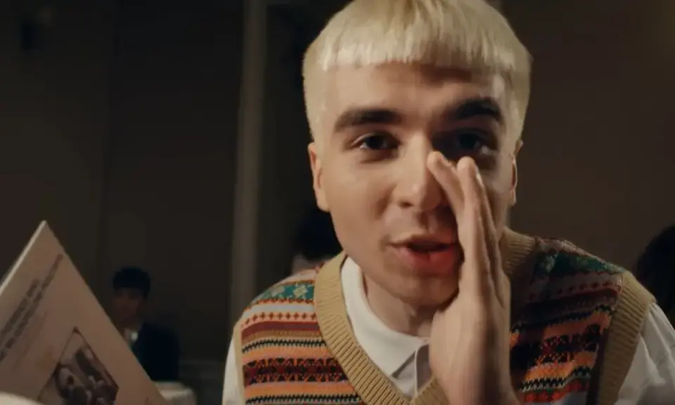 Młody mężczyzna z blond fryzurą, w kamizelce z kolorowym wzorem, trzyma rękę przy ustach, jakby coś szeptał. Obok niego leży blok mata tekst.
