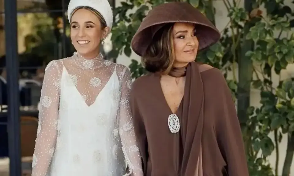 La madre de la novia, elegante con sombrero marrón, acompaña a la radiante novia con su vestido blanco adornado.