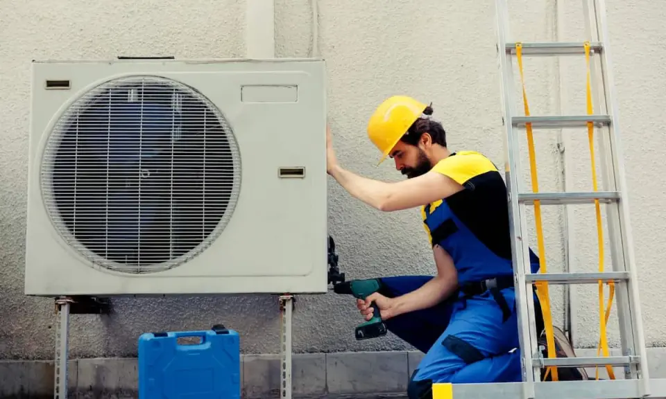 Technicien en bleu, casque jaune, installant une unité extérieure de pompe à chaleur. La garantie pompe à chaleur est assurée par ce professionnel.