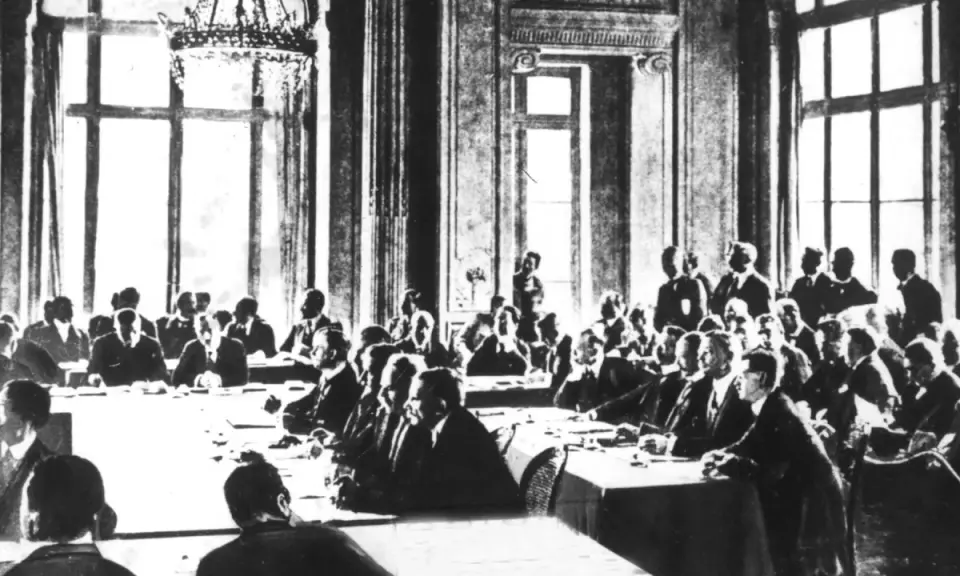 Männer in Anzügen sitzen an einem langen Tisch in einem prunkvollen Saal. Sie diskutieren die Bedingungen des Versailler Vertrags.
