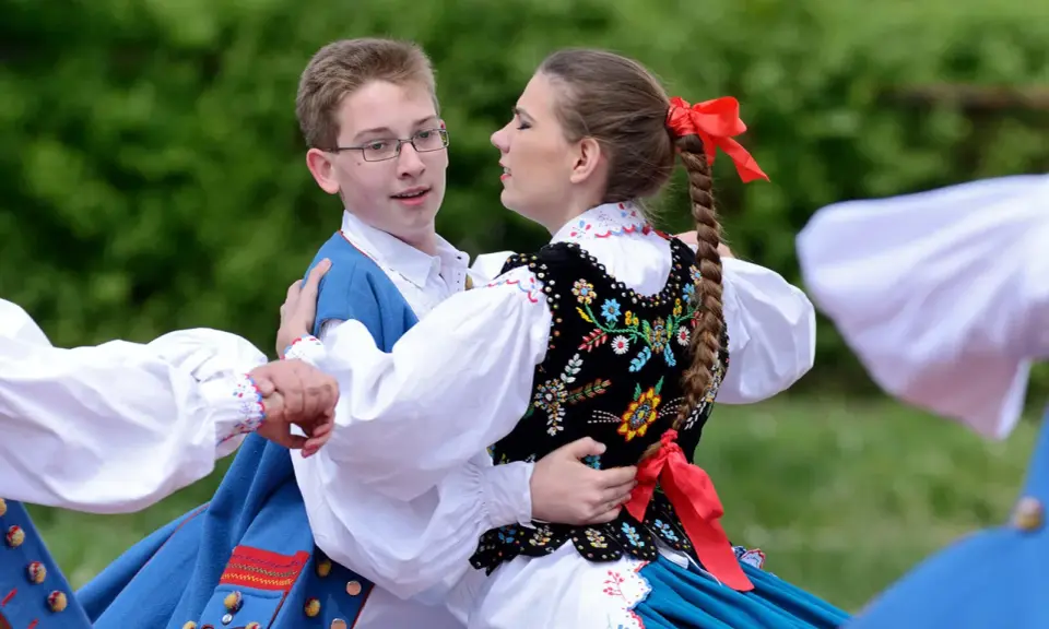 Młoda para w strojach ludowych tańczy mazura. Chłopiec w niebieskiej kamizelce, dziewczyna w haftowanej kamizelce i warkoczu z czerwoną wstążką.