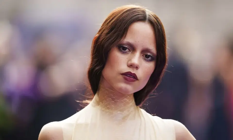 A young woman with auburn hair and a dramatic, dark lipstick looks directly at the camera. Her eye makeup is smudged, giving her a tired girl makeup look, as if she's been out all night.