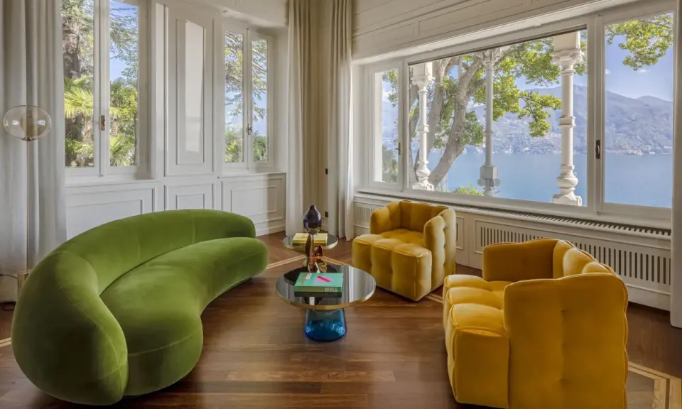 Salotto con divano verde curvo e poltrone gialle, tavolini di design e vista lago. Un'elegante casa Art Déco.