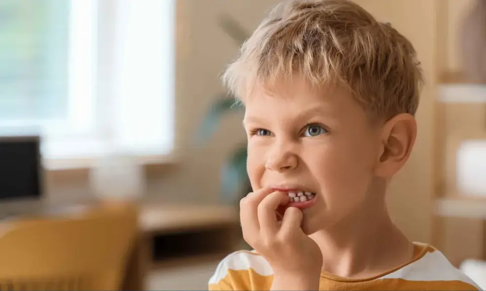 Chłopiec z blond włosami i niebieskimi oczami, który gryzie paznokcie. Może to być objaw stresu lub zespołu Tourette'a.