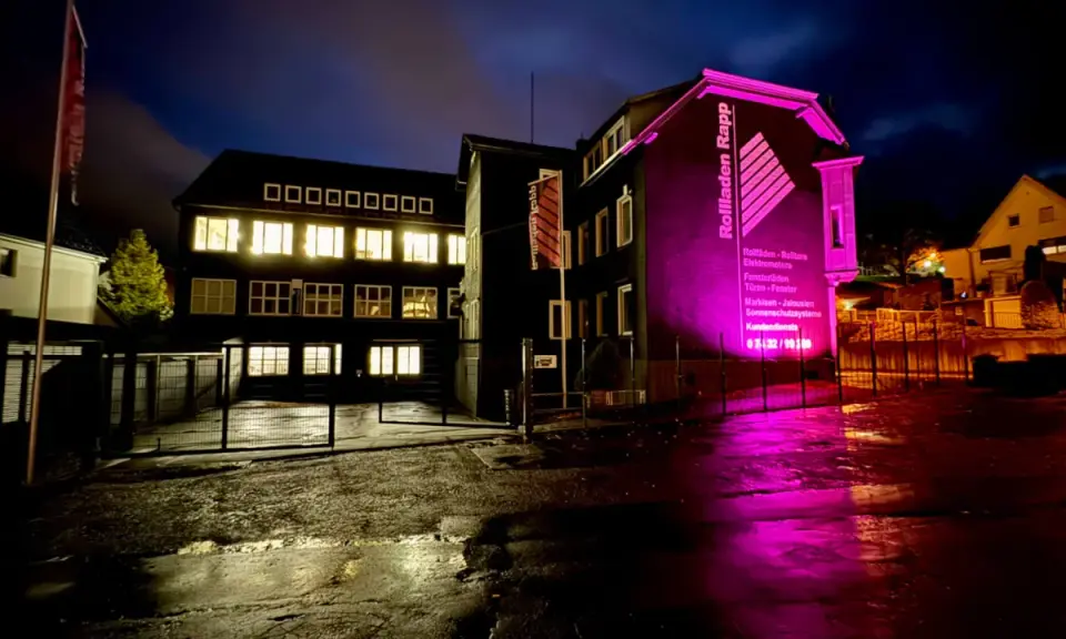 Gebäude mit leuchtenden Fenstern und pinker Beleuchtung. "Rolladen Rapp" wirbt für Nothandkurbel Rolladen, Pflicht für Sicherheit.