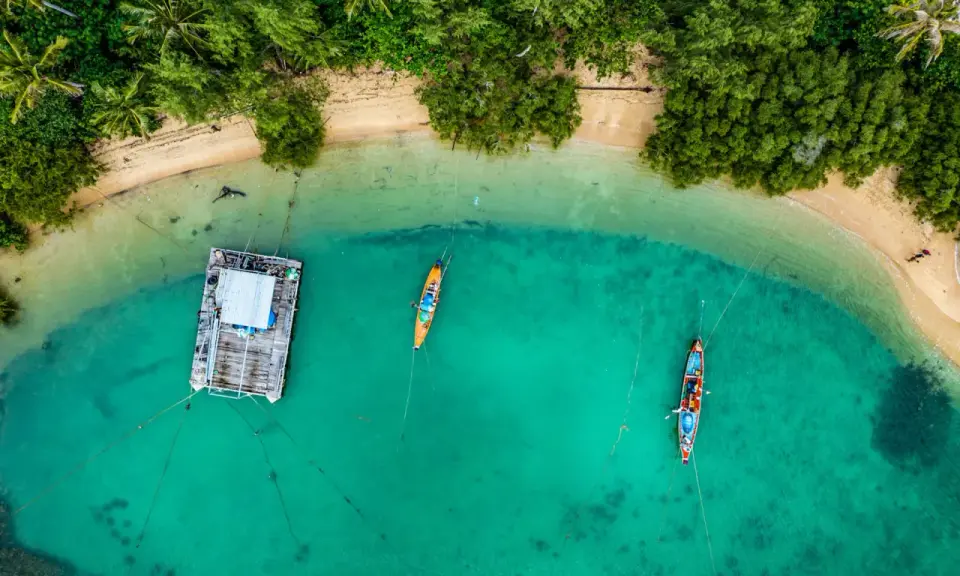 Drohne über Thailand: Ein Blick von oben auf eine schwimmende Plattform und zwei Boote im türkisfarbenen Meer, umgeben von üppiger Vegetation und Sandstrand.