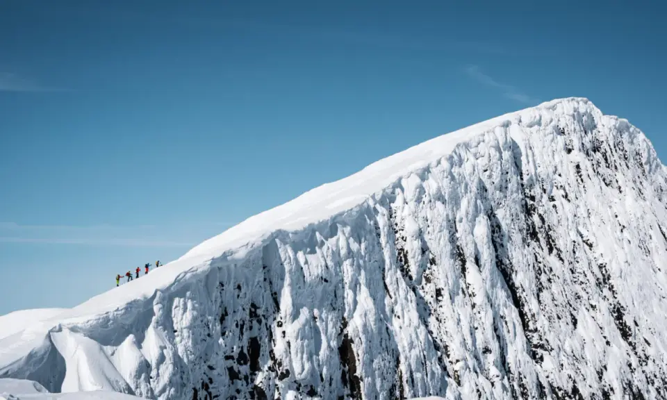 En grupp skidåkare på en helags topptur, med en dramatisk snöklädd bergssida och klarblå himmel.