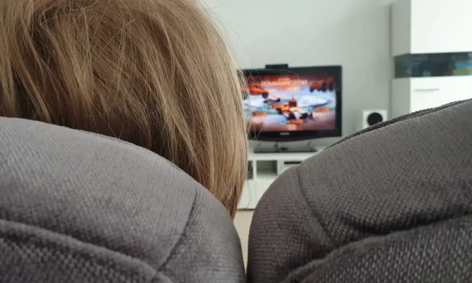 Niemowlę z blond włosami ogląda bajkę na telewizorze w tle, siedząc na kanapie.