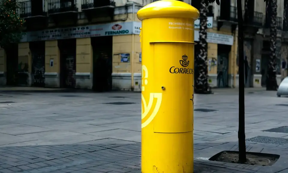Jasnożółta skrzynka pocztowa Correos, symbol poczty Hiszpanii, stoi na brukowanym placu przed budynkami.