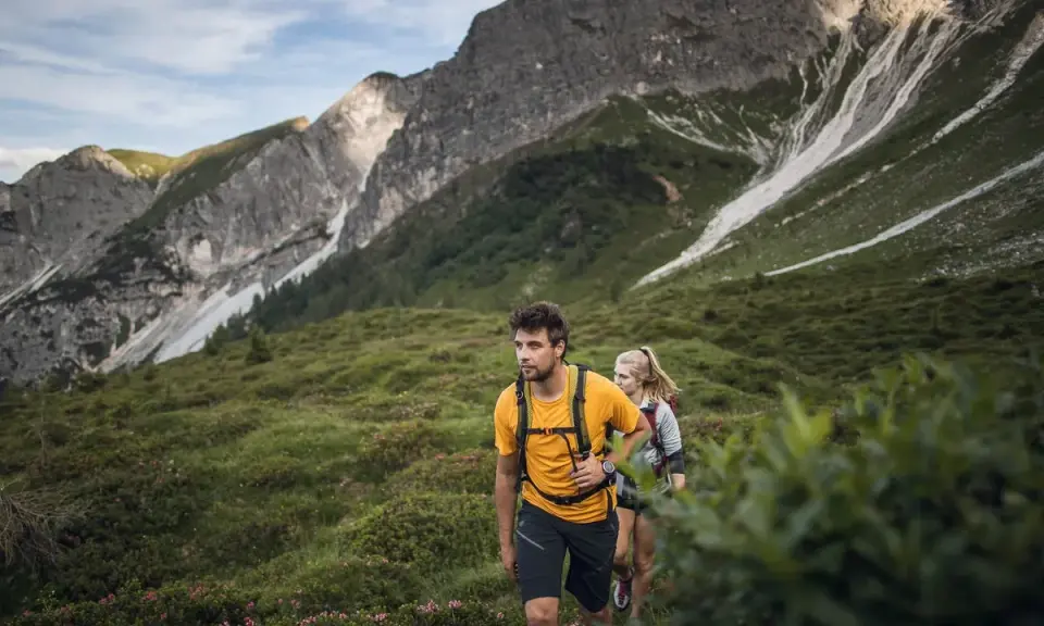 Coppia in escursioni alto adige, camminano su un sentiero verdeggiante con montagne imponenti sullo sfondo.