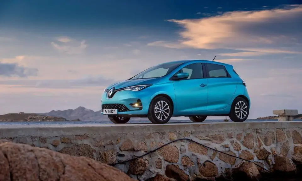 Un'elegante Renault Zoe blu, uno dei tanti modelli Renault, è parcheggiata su un muretto di pietra con il mare e le montagne sullo sfondo.