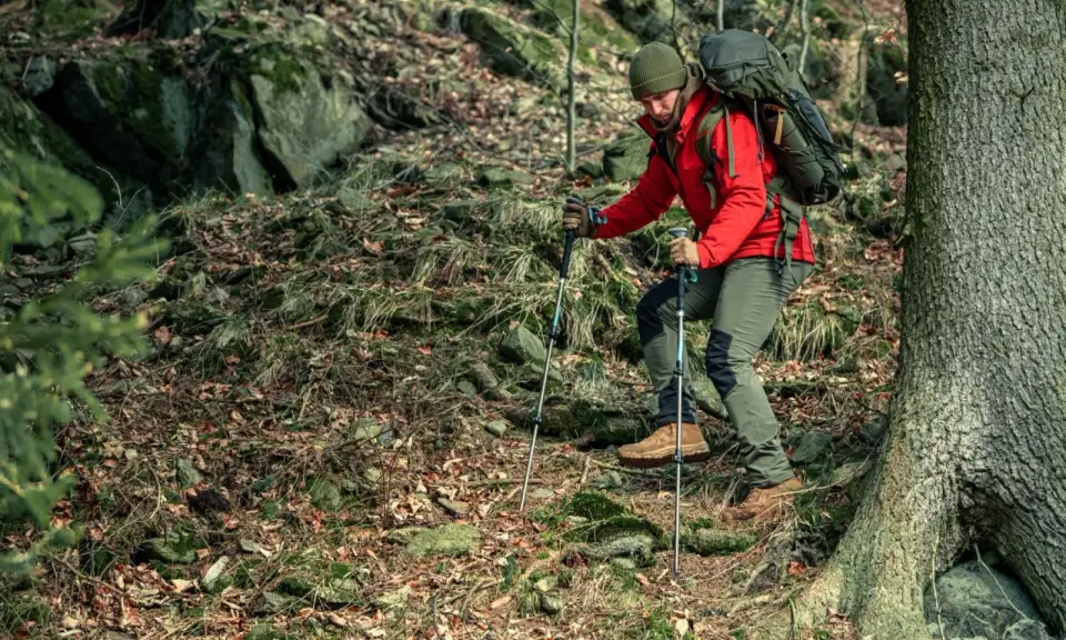 Mężczyzna w czerwonej kurtce i zielonej czapce wspina się po leśnym zboczu, używając kijków trekkingowych. To właśnie **outdoor** – życie na świeżym powietrzu.