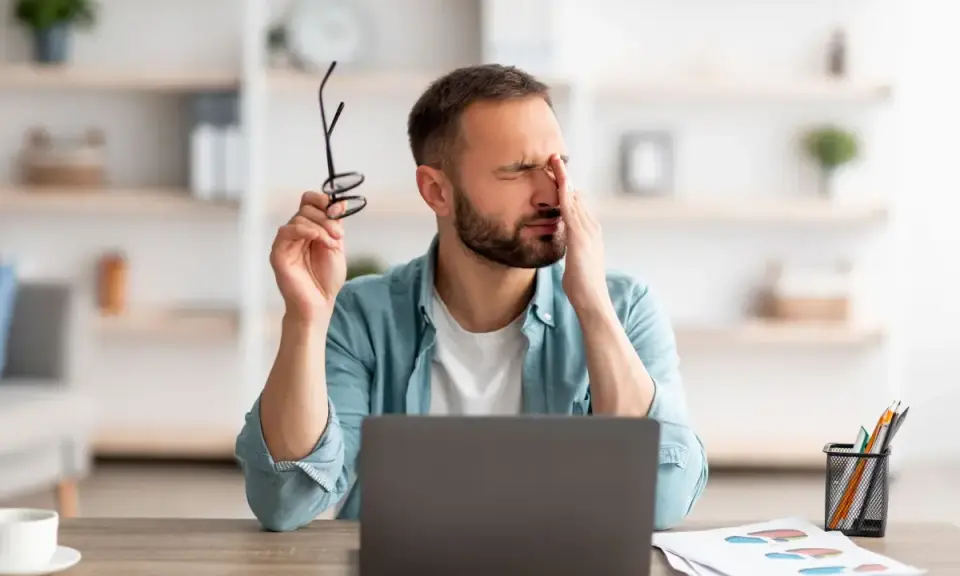 Homme fatigué, se frottant les yeux, lunettes à la main, devant son ordinateur. Il souffre de mal aux yeux à cause des écrans.