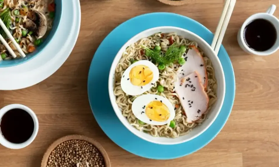 Miseczka z tantanmen, makaronem, jajkiem na twardo, plastrami mięsa i zielonym koperkiem. Obok sos sojowy i nasiona sezamu.