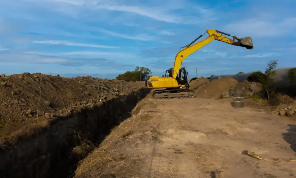 Excavadora amarilla trabajando en la creación de varios tipos de zanjas, con montones de tierra y tuberías grandes en el fondo.
