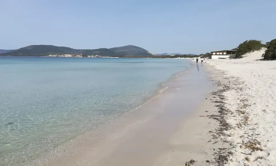 Spiaggia della Fertilia con acque cristalline, sabbia bianca e un edificio in lontananza. Due persone passeggiano lungo la riva.