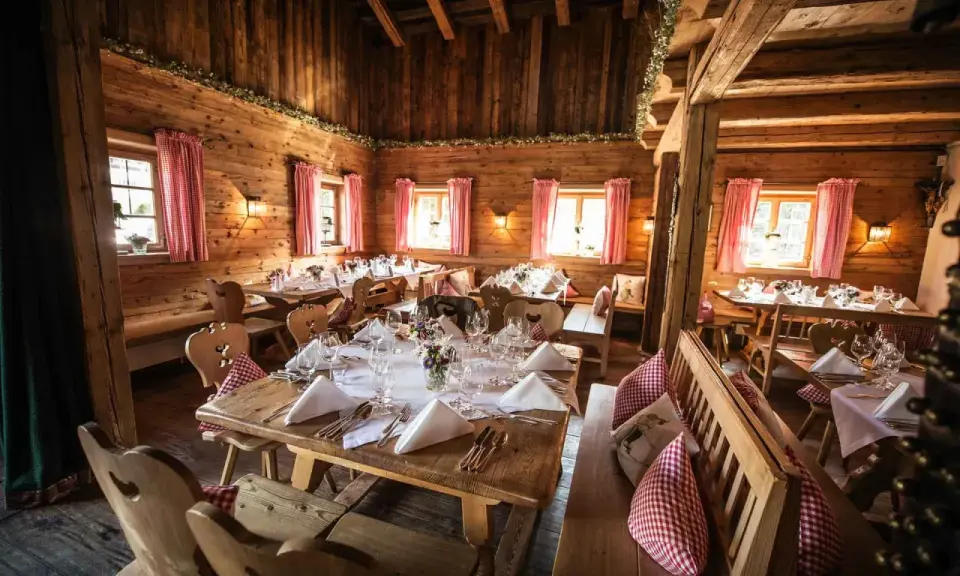 Gemütliche Holzhütte, festlich gedeckte Tische für eine Egerner Alm Hochzeit. Rote Karovorhänge und Blumen schmücken den rustikalen Raum.