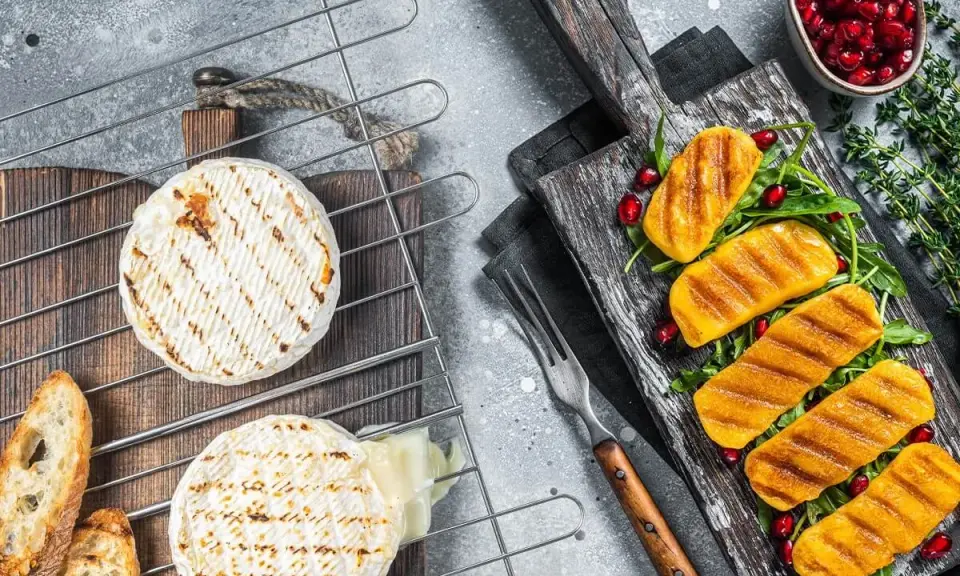 Dwa okrągłe sery camembert grillowane na ruszcie, obok pieczywo i grillowane kawałki sera na desce z granatem.
