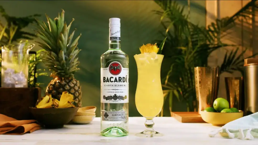 Butelka białego rumu Bacardi obok koktajlu Piña Colada, z ananasami i limonkami w tle.