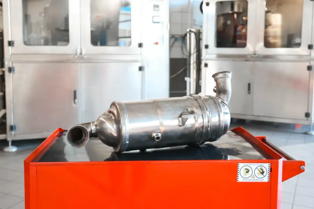 Metalowy element układu wydechowego, prawdopodobnie gpf, leży na pomarańczowej skrzyni. W tle widać stalowe szafy.