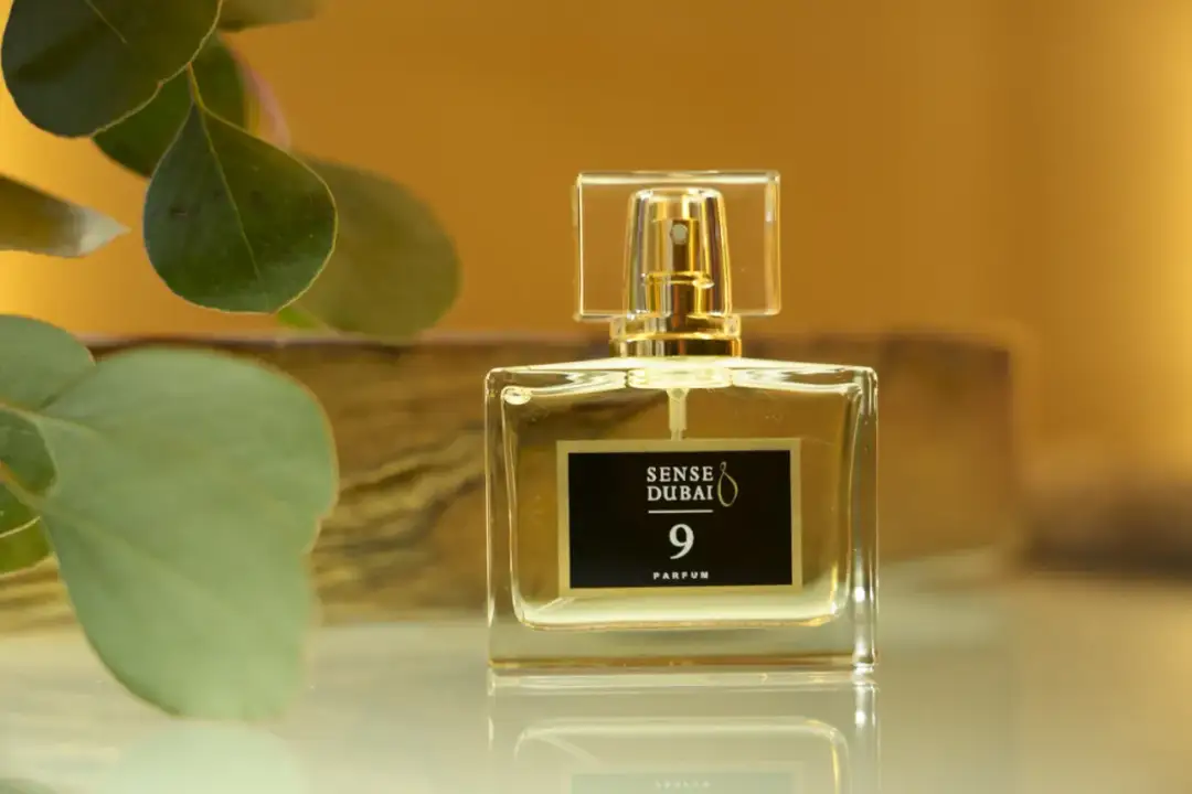 Eleganckie perfumy damskie z Dubaju, Sense Dubai 9 Parfum, w szklanym flakonie z złotymi detalami, otoczone zielonymi liśćmi.