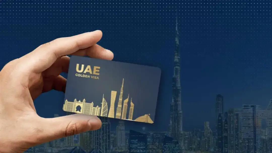 Dłoń trzyma kartę "UAE GOLDEN VISA" na tle panoramy Dubaju z Burdż Chalifą.