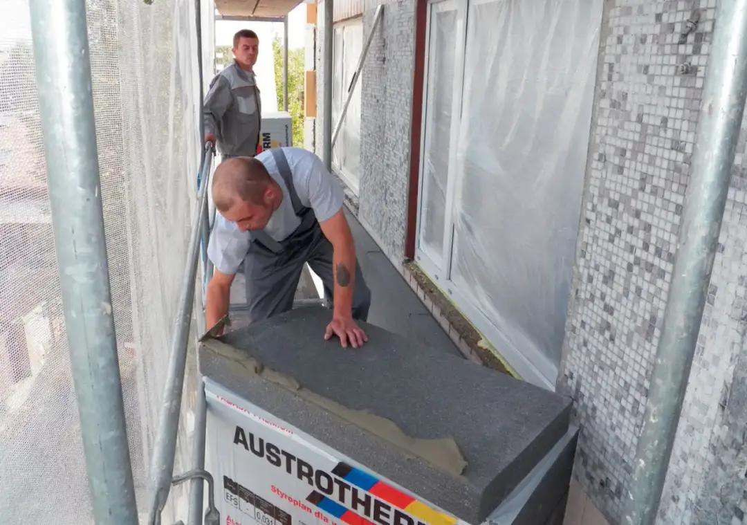 Budowlaniec układa styropian pod wylewkę na balkonie. Obok stoi drugi pracownik.