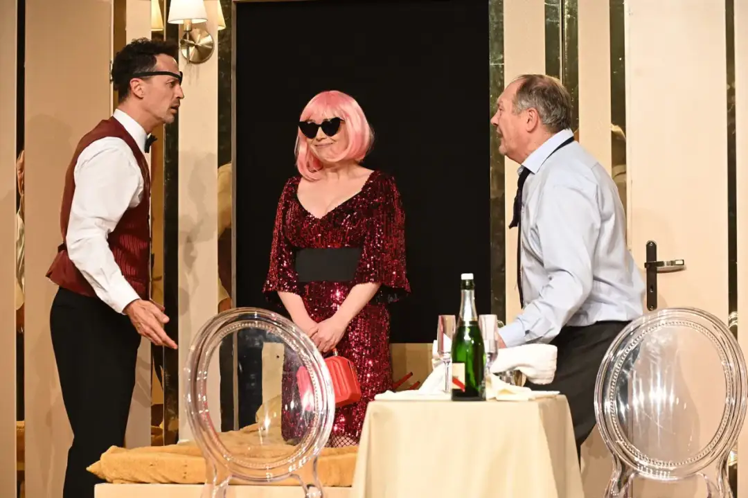 Trzech aktorów na scenie Teatru Capitol Kraków. Kobieta w błyszczącej sukni i peruce, mężczyźni w eleganckich strojach.