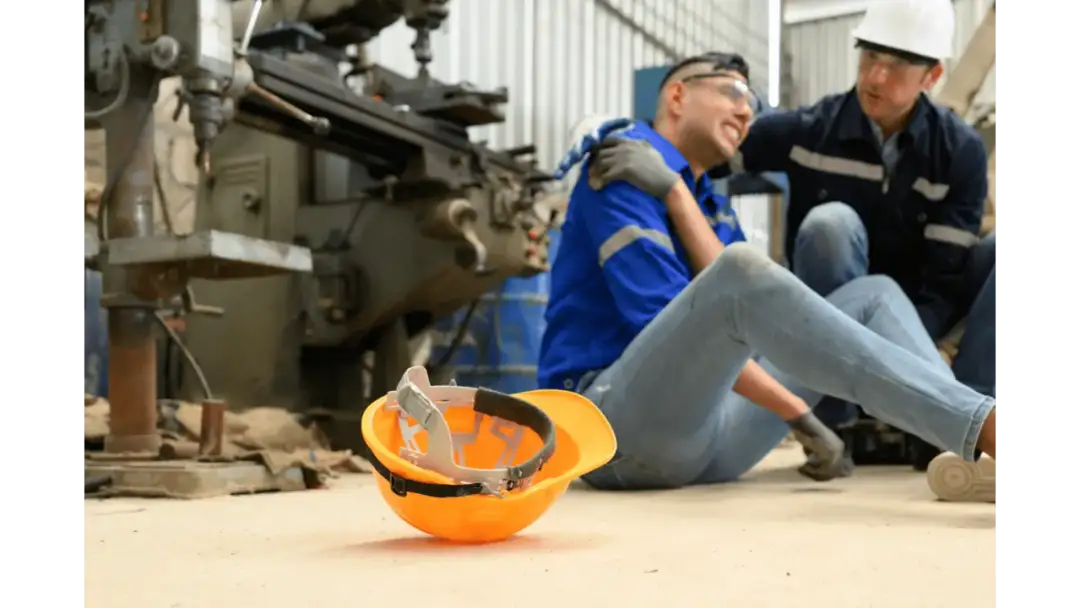 Praca na czarno konsekwencje dla pracownika w razie wypadku: mężczyzna po urazie siedzi na ziemi, kolega mu pomaga. Pomarańczowy kask leży obok.