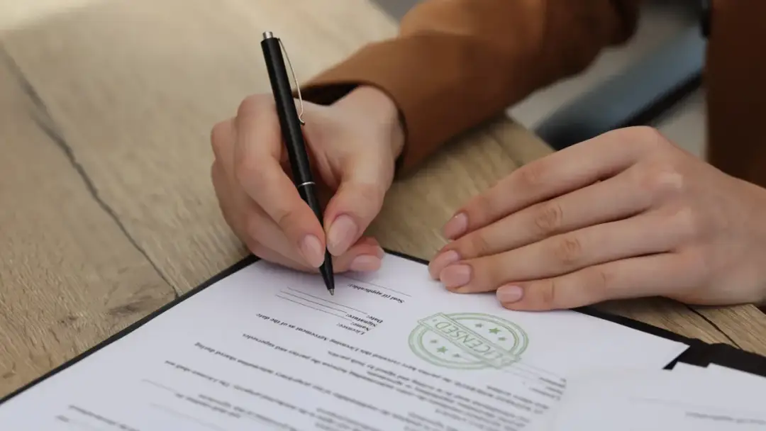 Dłoń podpisuje dokument, na którym widnieje pieczęć "LICENSED". To ważny krok do uzyskania licencji.