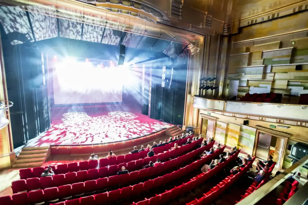Widownia Teatru Capitol wypełniona widzami, siedzącymi w czerwonych fotelach. Na scenie grają światła, tworząc magiczną atmosferę.