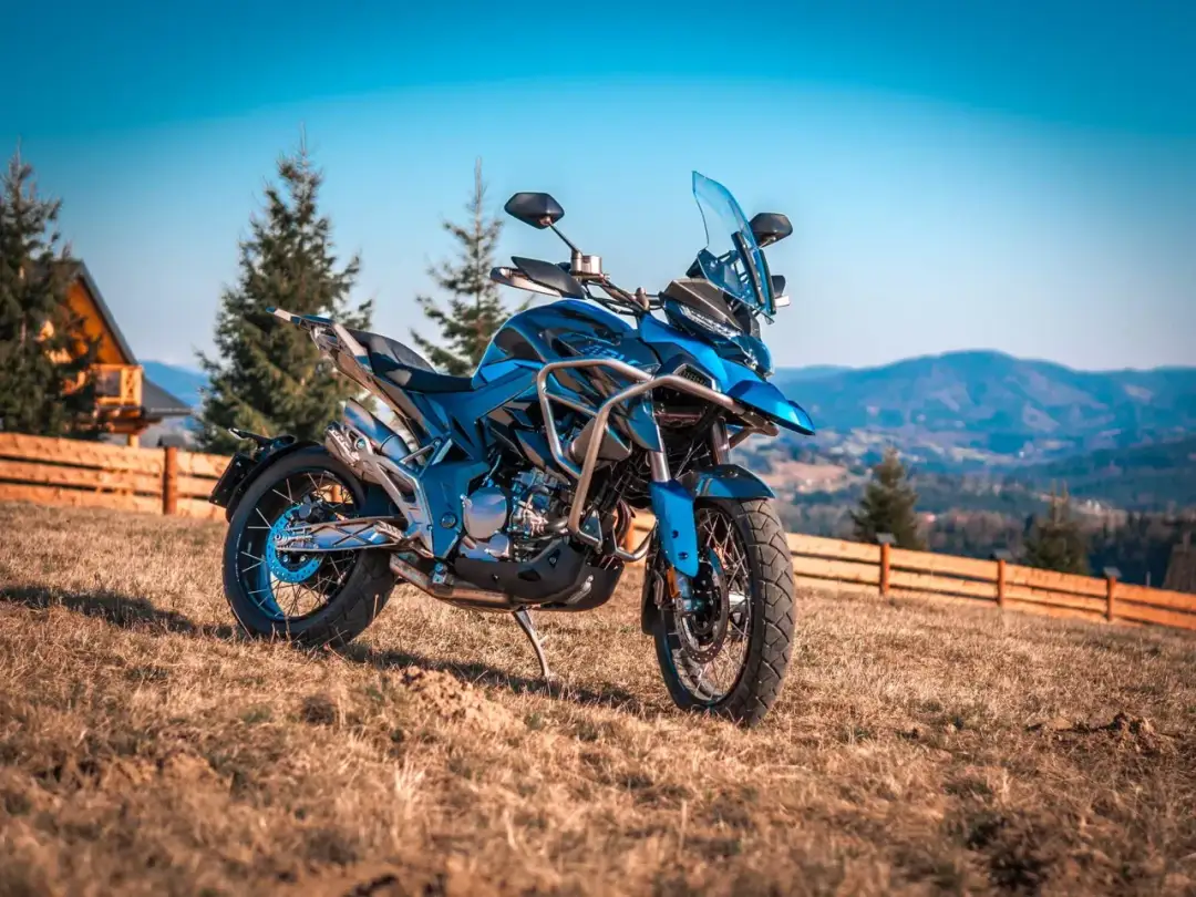 Niebieski motocykl turystyczny na trawiastym wzgórzu, z widokiem na góry i las.