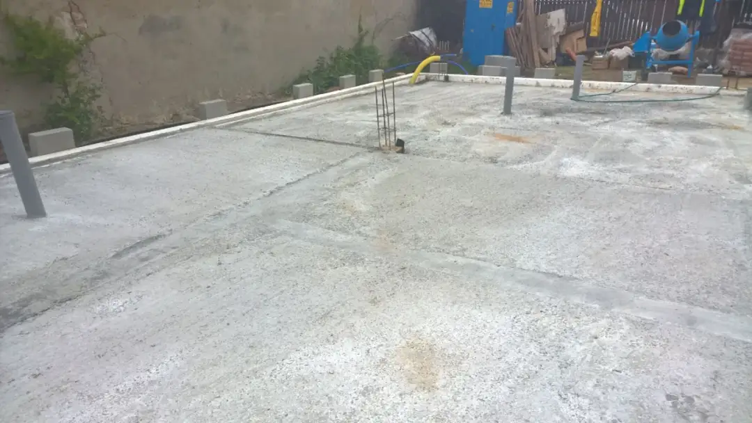 Plac budowy z nową wylewką betonową, gdzie widać, jak połączyć stary beton z nowym. W tle betonowa ściana i ogrodzenie.