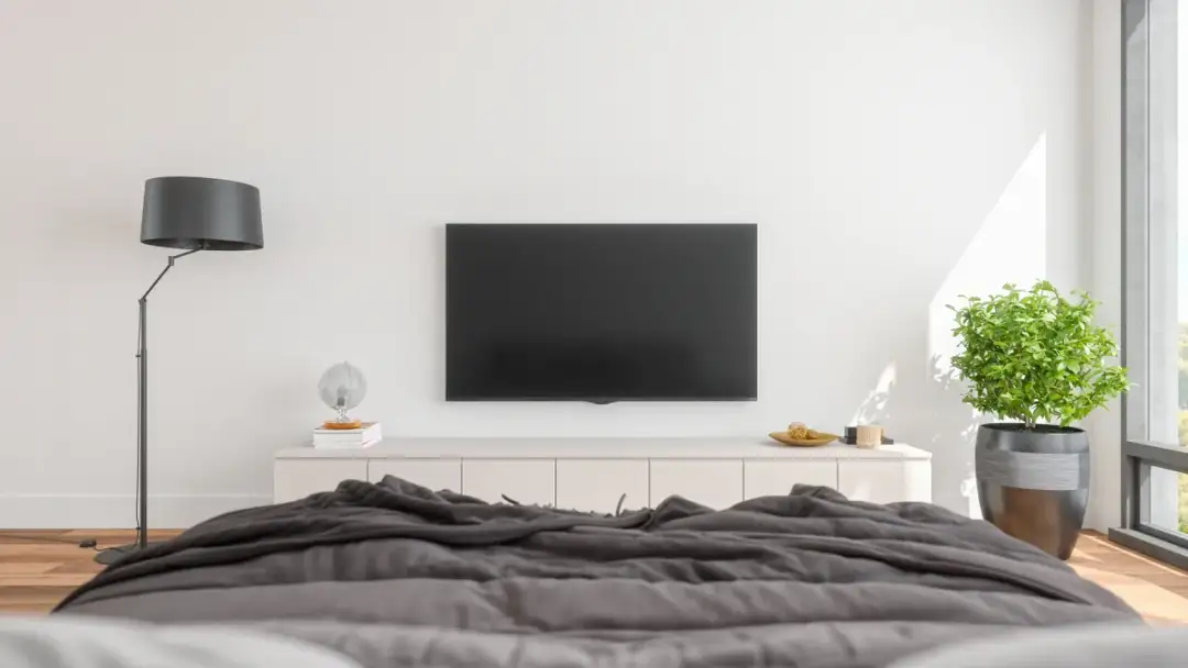 Nowoczesny salon z TV na ścianie, idealna wysokość od podłogi. Obok lampa, komoda z dekoracjami i roślina.