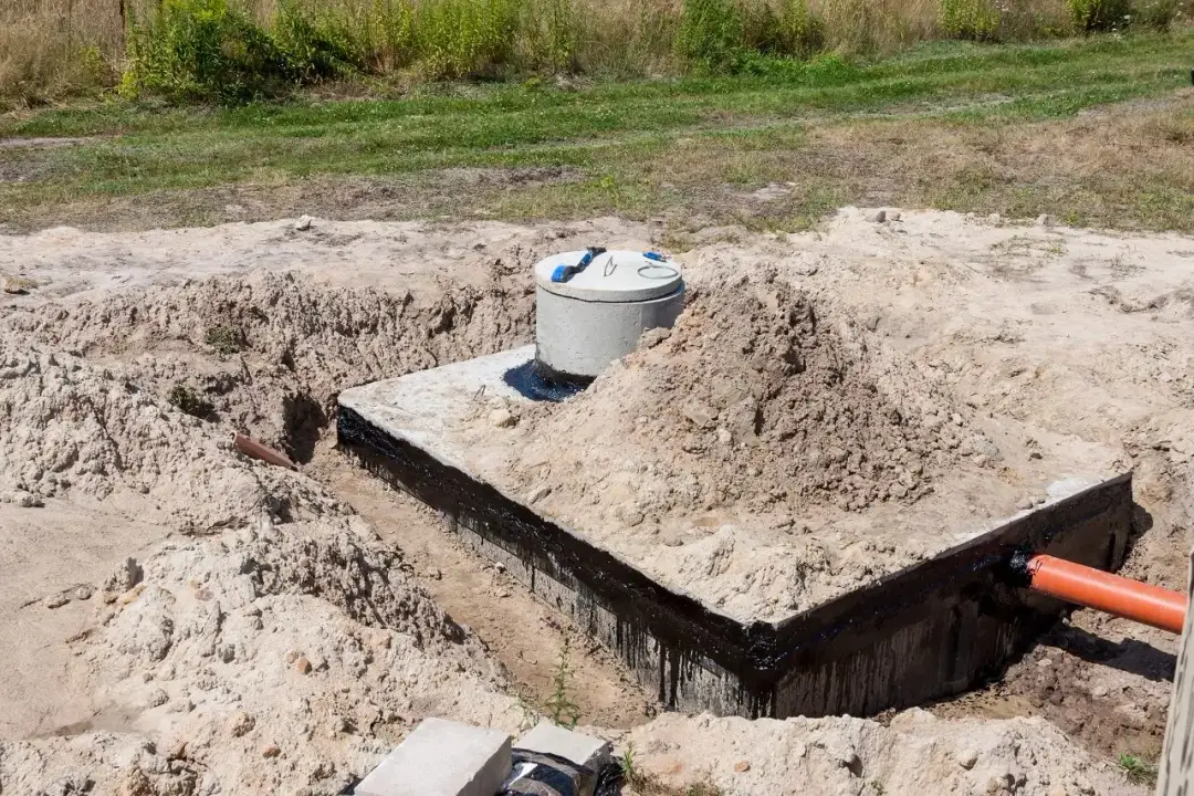 Betonowe szambo w wykopie, z widoczną izolacją i rurą odpływową.