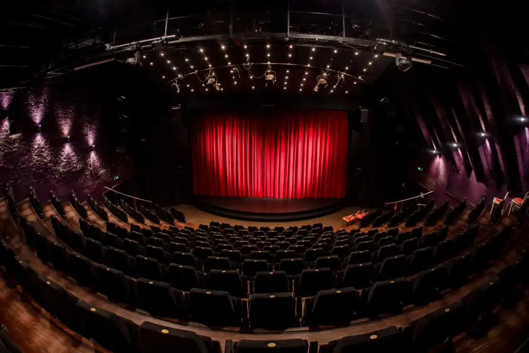 Pusta sala teatralna z czerwonymi kurtynami i rzędami czarnych foteli. Czekamy na spektakl z repertuaru Teatru Janda.