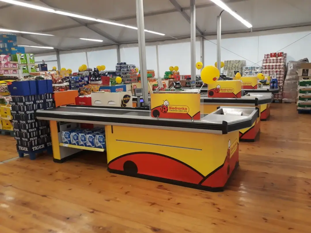 Kasy w Biedronce, gdzie pracownik zarabia na codzienne niskie ceny. W tle półki z produktami i żółte balony.