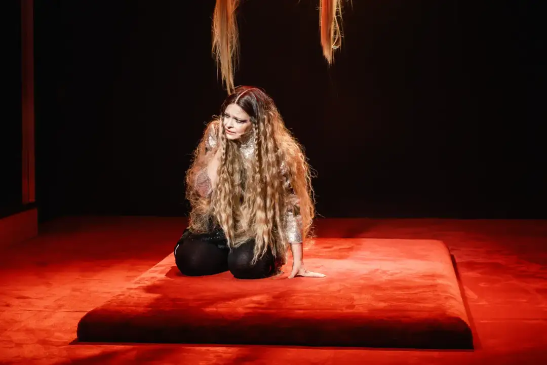 Aktorka na czerwonej scenie w teatrze w Warszawie, w błyszczącej kreacji i z długimi włosami, przeżywa dramatyczny moment.