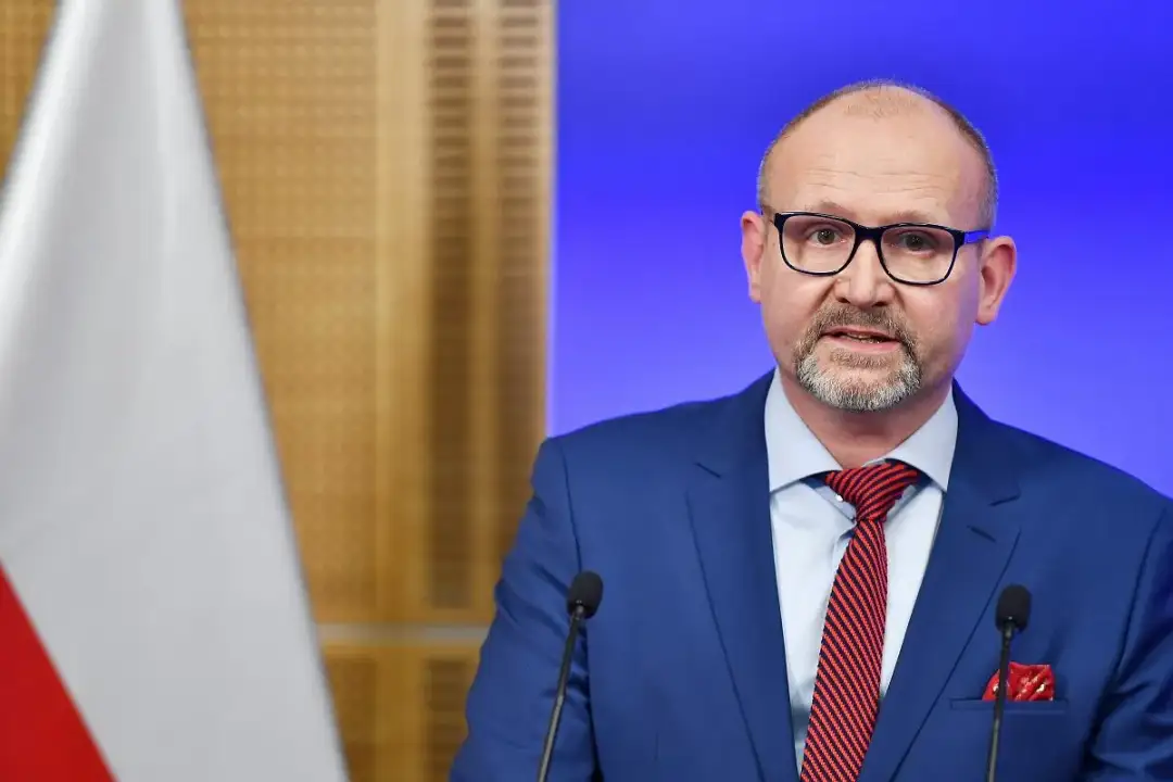 Prokurator krajowy w garniturze i okularach przemawia przy mikrofonie, obok polskiej flagi.