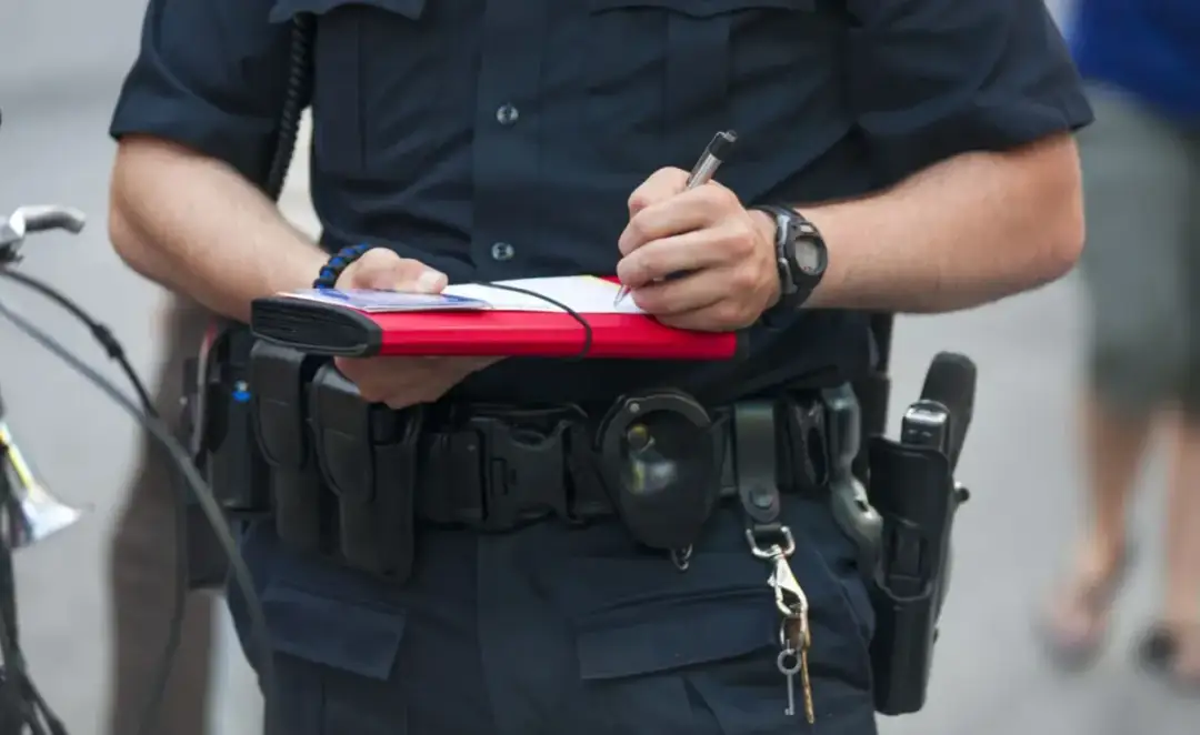 Policjant spisuje mandat, być może według taryfikatora mandatów 2025. Na pasie ma kaburę z bronią i kajdanki.