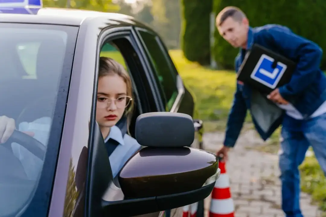 Dziewczyna za kierownicą, zdenerwowana, czeka na egzaminatora z teczką "L". Tak wygląda egzamin na prawo jazdy.