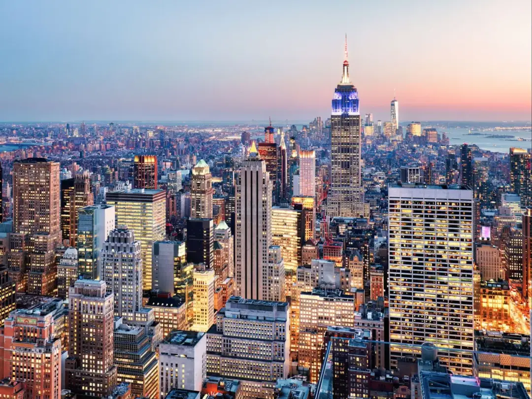Widok na rozświetlony Nowy Jork o zmierzchu, z Empire State Building w centrum. Zastanawiasz się, ile kosztuje lot do USA?