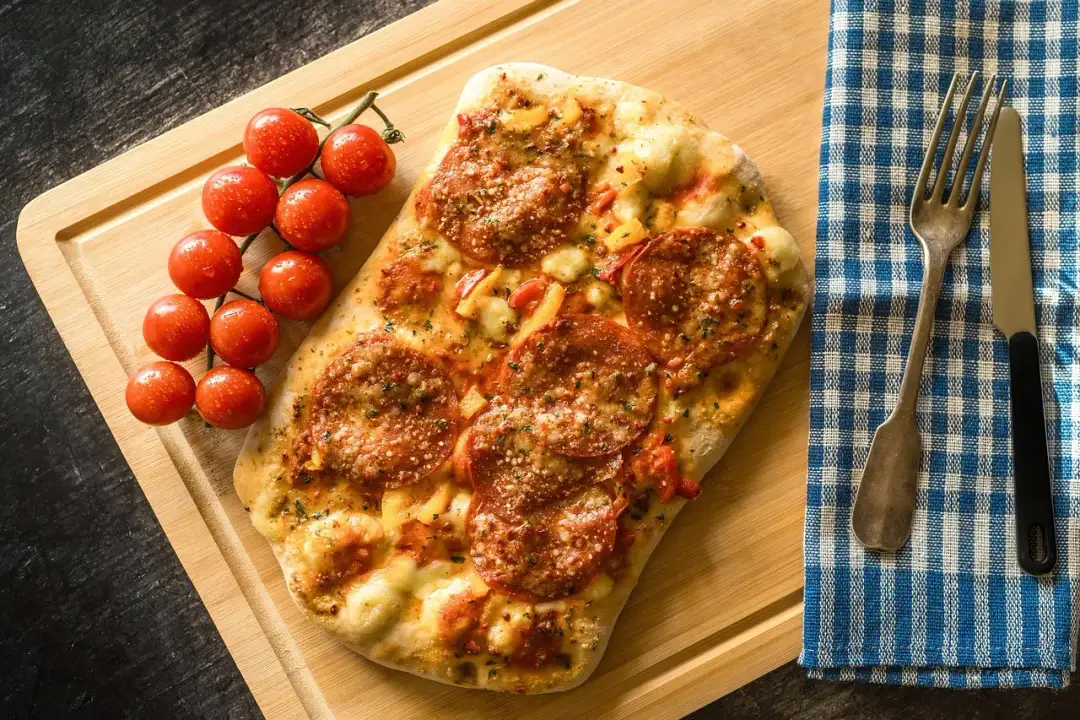 Pizza pepperoni z pomidorkami koktajlowymi na desce, obok serwetka i sztućce.