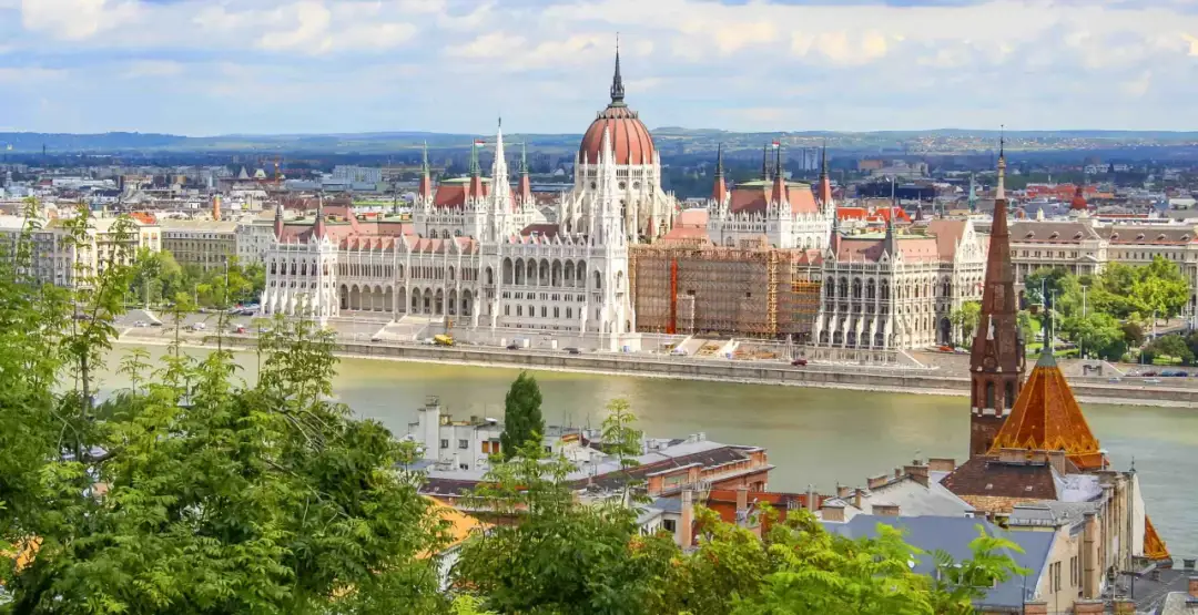 Widok na Parlament w Budapeszcie, cel podróży pociągiem do Budapesztu z Polski. Rzeka Dunaj i zielone drzewa na pierwszym planie.