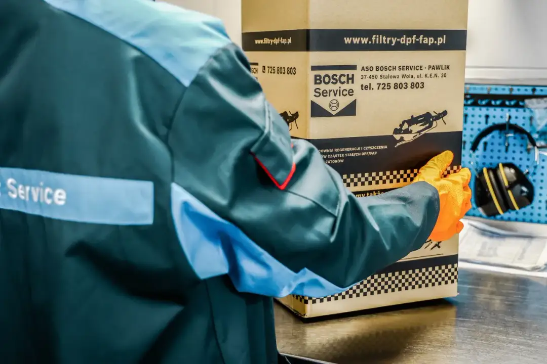 Pracownik w pomarańczowych rękawiczkach trzyma karton z logo Bosch Service. Dowiedz się, ile trwa wypalanie DPF na postoju.