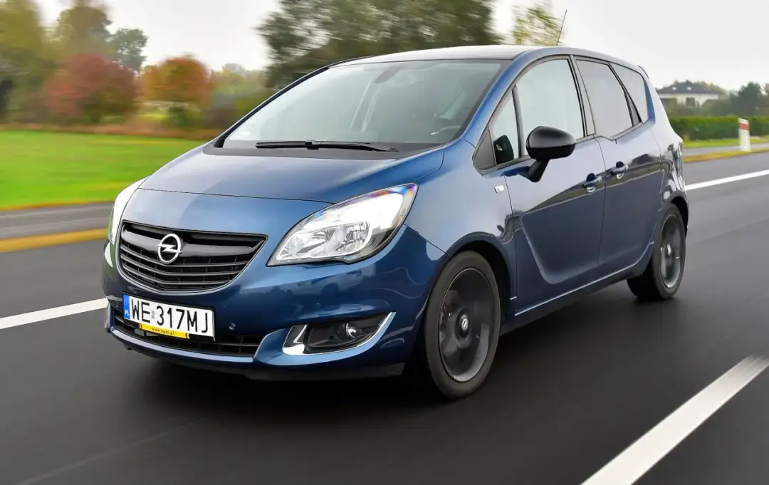 Niebieski Opel Meriva B 1.4 Turbo pędzi po drodze, otoczony jesiennym krajobrazem.