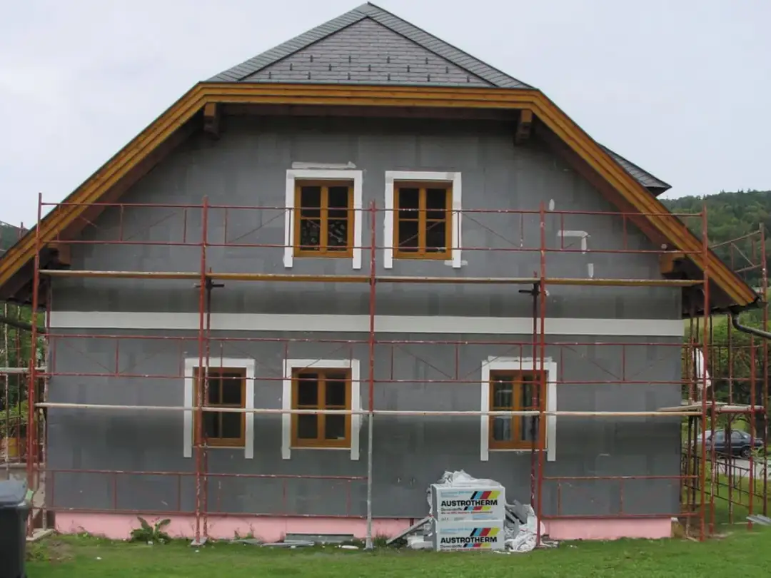 Dom w trakcie ocieplania. Szara fasada, drewniane okna, rusztowanie. Materiały budowlane Austrotherm wskazują na dbałość o niski współczynnik przewodzenia ciepła.