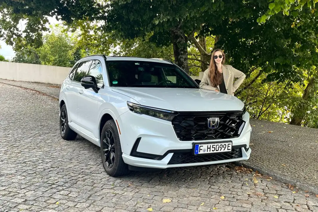 Uśmiechnięta kobieta opiera się o białą Honda CRV 2025 na brukowanej uliczce.