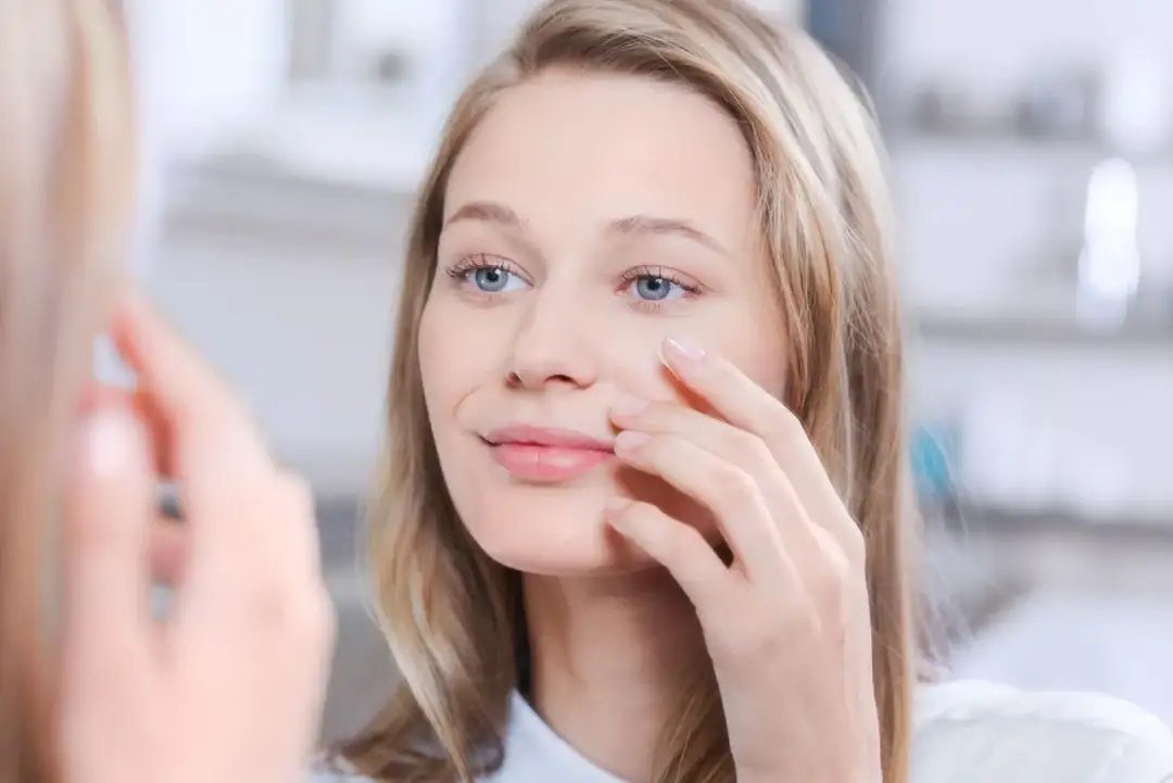 Delikatny meszek na twarzy młodej kobiety, która dotyka policzka, patrząc w lustro.