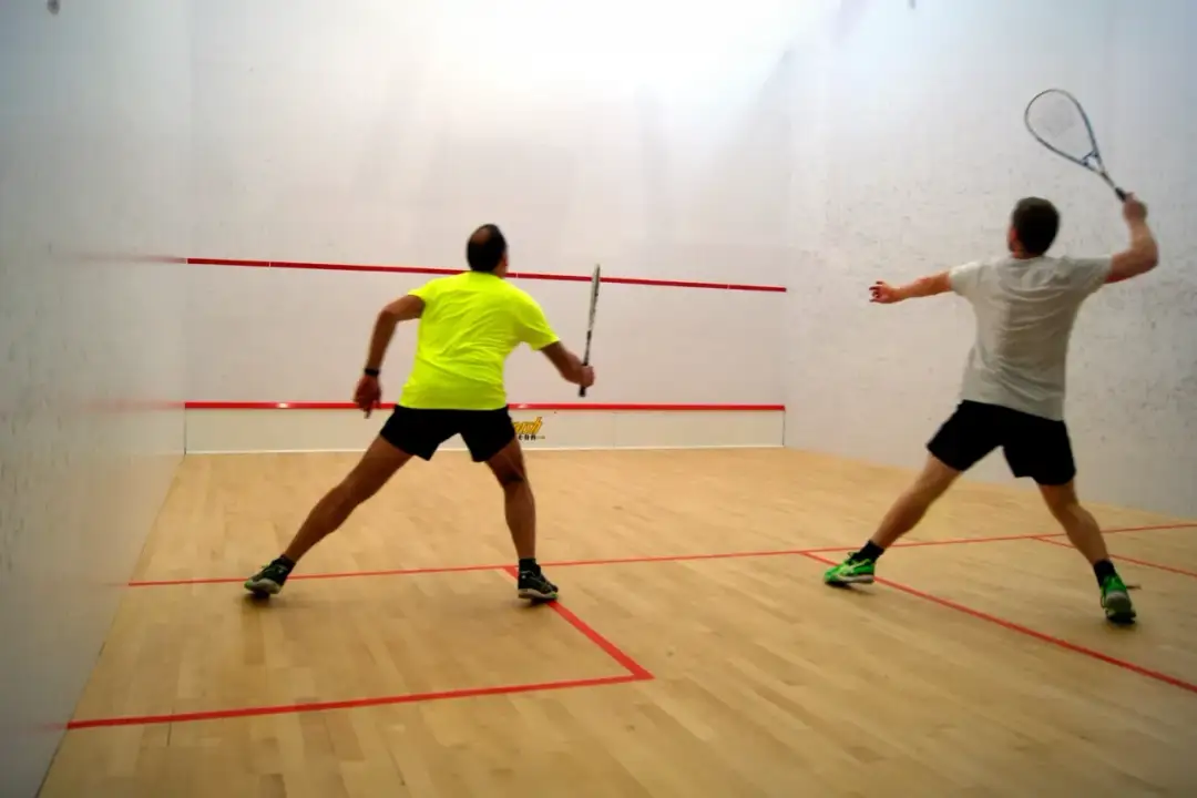 Dwóch mężczyzn gra w squasha na boisku z drewnianą podłogą i białymi ścianami.